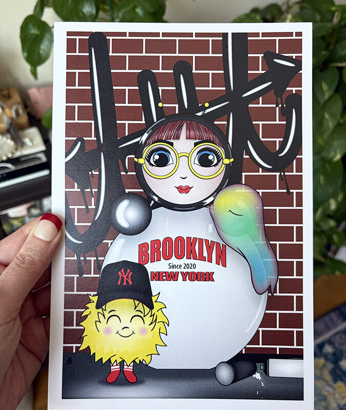 Brooklyn graffiti mini print held in hand, featuring O with yellow glasses and EM in a Yankees hat against a brick wall with black graffiti.