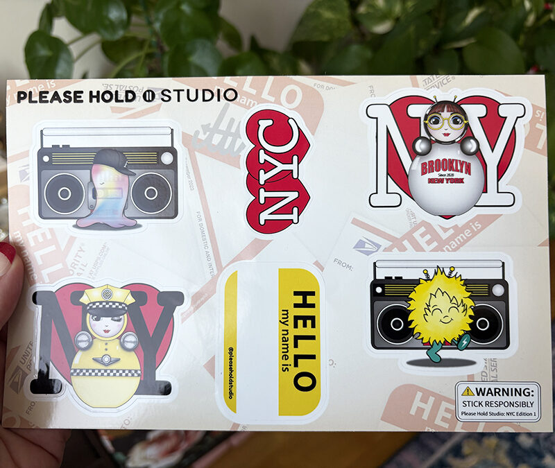 NYC Edition 1 sticker sheet held in hand, featuring O, EM, a boombox, taxi O, and “Hello My Name Is” designs.