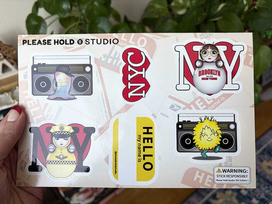 NYC Edition 1 sticker sheet held in hand, featuring O, EM, a boombox, taxi O, and “Hello My Name Is” designs.