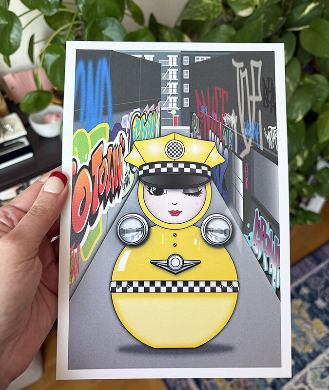 Taxi O mini art print held in hand, featuring the yellow taxi-inspired O character in a graffiti-covered NYC alley.