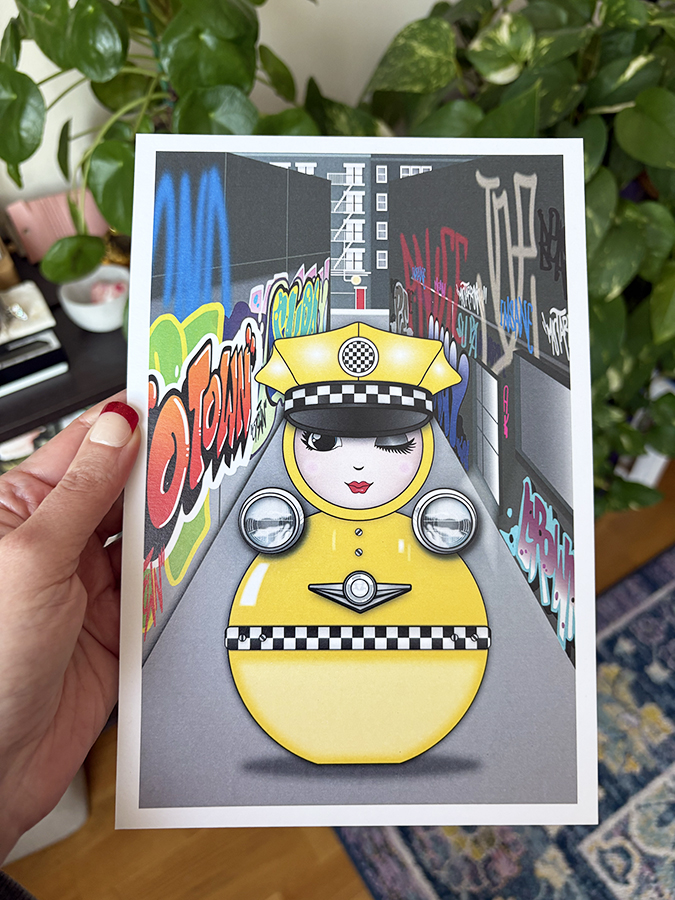 Taxi O mini art print held in hand, featuring the yellow taxi-inspired O character in a graffiti-covered NYC alley.